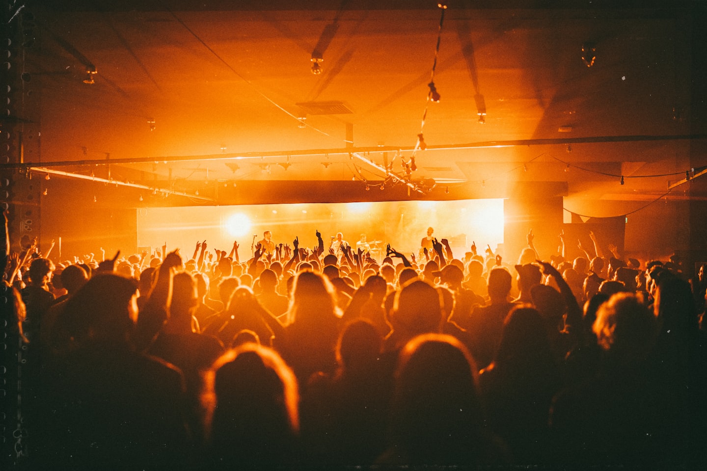 Crowd with hands raised enjoys a band performing on a brightly lit stage.