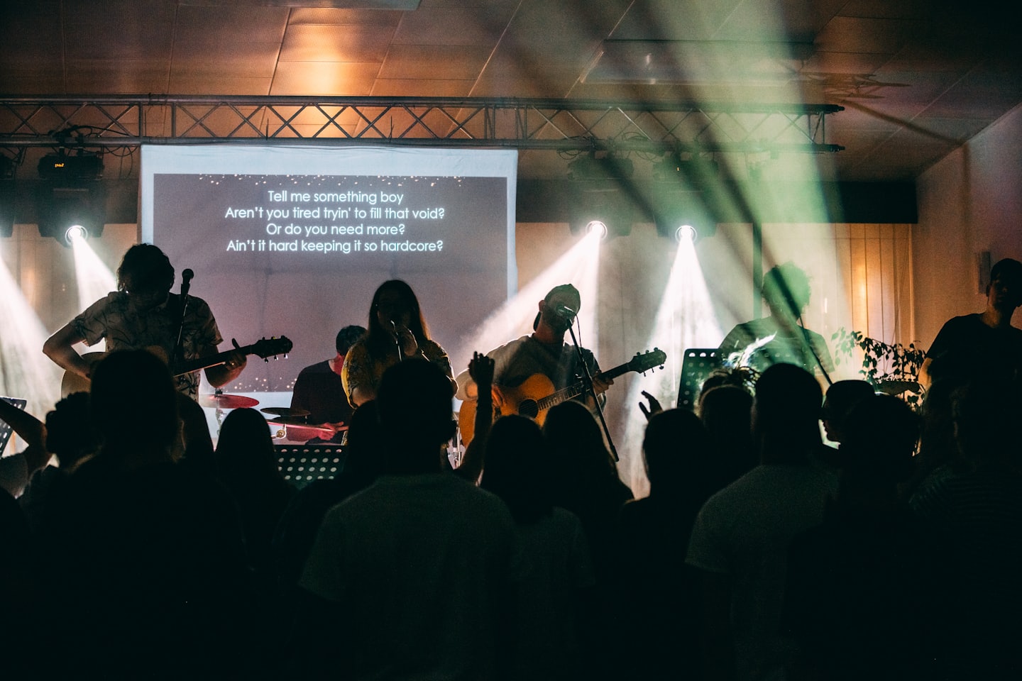Musicians perform on a brightly lit concert stage with lyrics on screen.